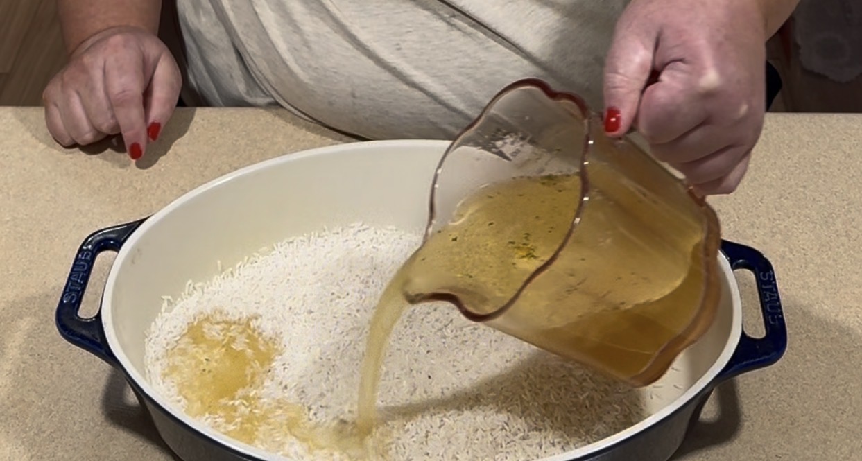 Hands pouring golden liquid from a measuring pitcher into a white baking dish filled with raw rice.