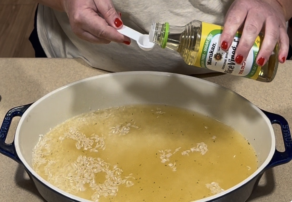 A person pours rice vinegar from a bottle into a small measuring spoon over a pot of rice and liquid.