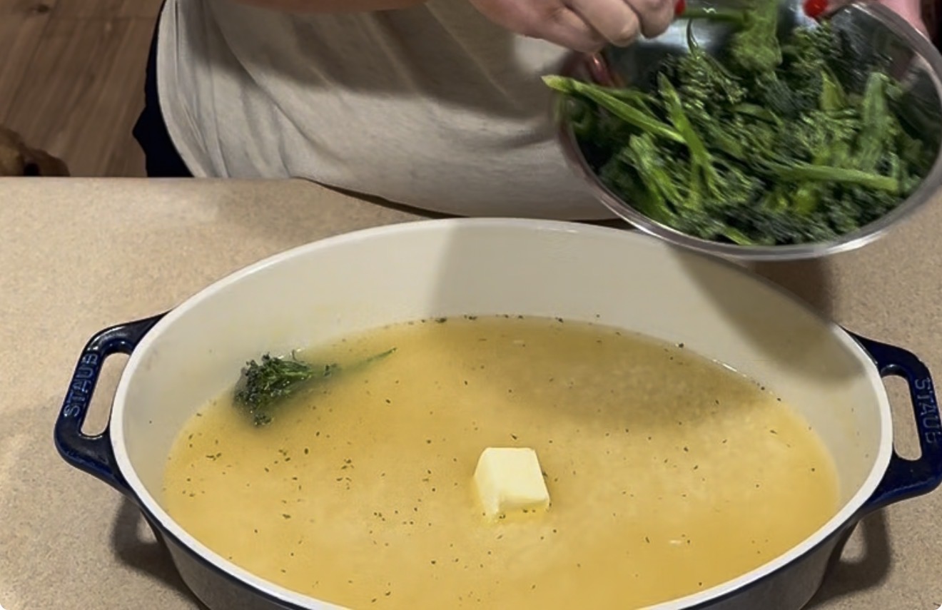 A person pours a bowl of broccolini into a wide dish containing melted butter and seasoned liquid.