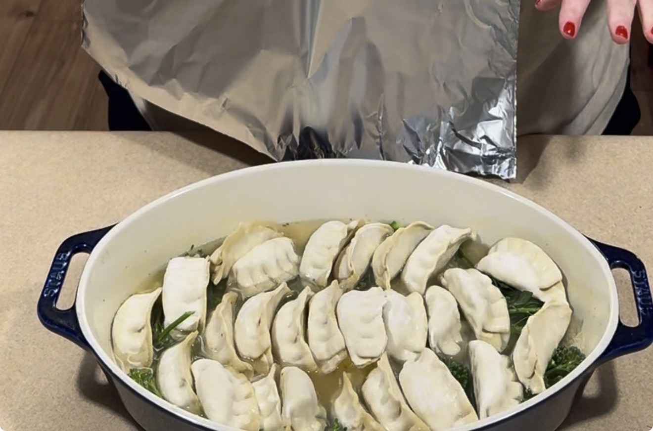A white oval baking dish filled with dumplings and broccoli in broth, with a sheet of aluminum foil held above it.