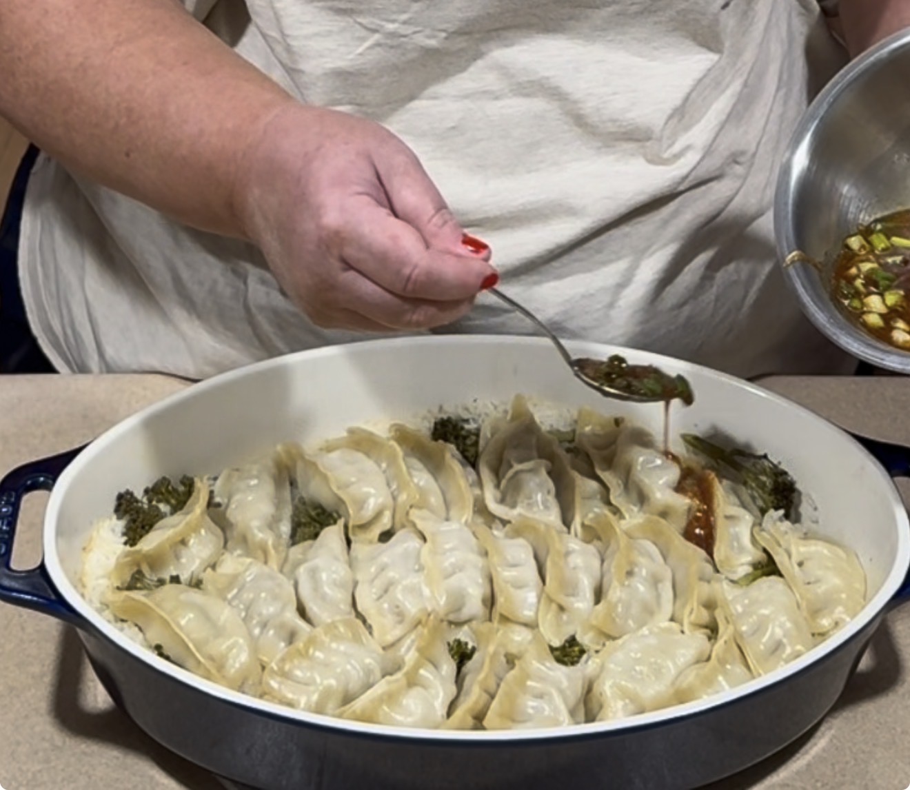 A person spoons sauce over a dish of dumplings and broccoli arranged in a shallow baking pan.