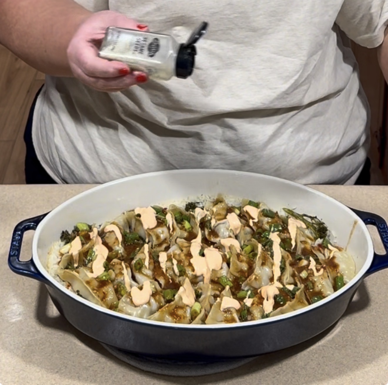 A person shakes sesame seeds over a casserole dish filled with dumplings drizzled with sauce.