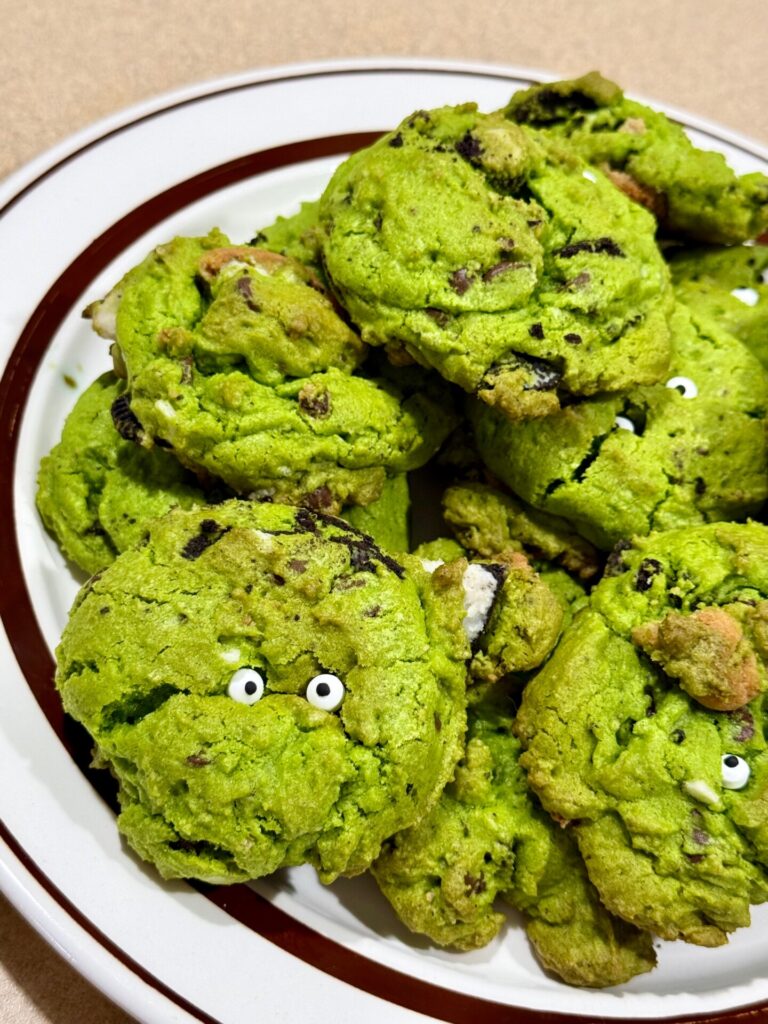 A pile of bright green cookies with chocolate chips and edible candy eyes on a white plate with a brown rim.