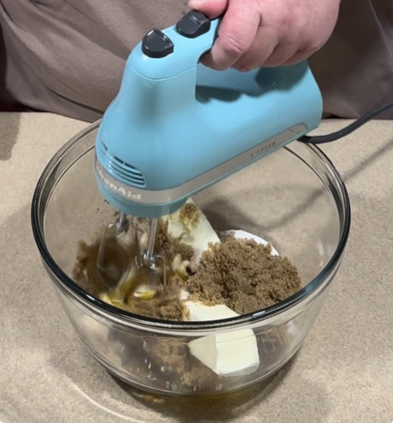 A hand uses a light blue handheld electric mixer to blend butter and brown sugar in a glass bowl.