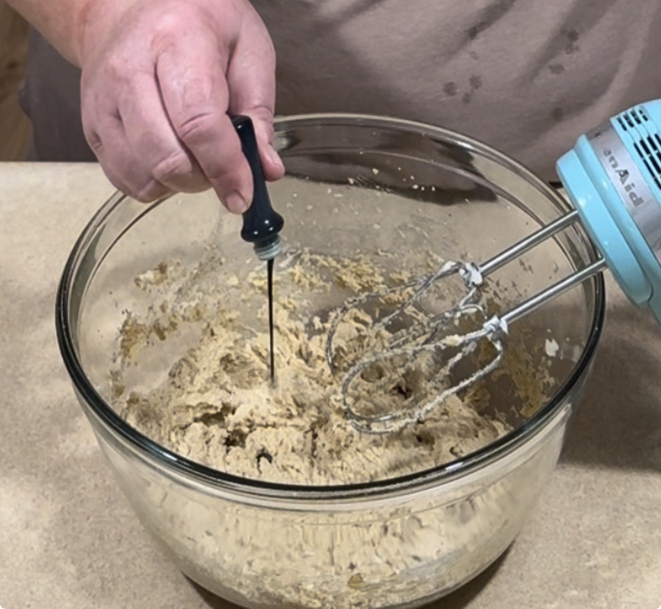 A hand adds liquid from a small black bottle into a glass mixing bowl containing cookie dough next to a hand mixer.