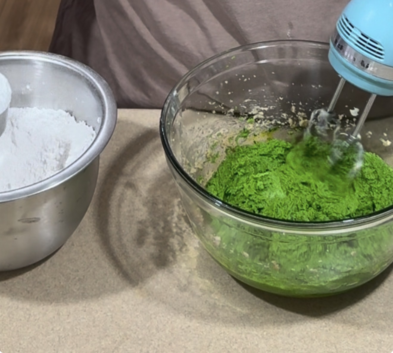 A hand mixer blends vibrant green batter in a glass bowl, next to a metal bowl containing powdered sugar.