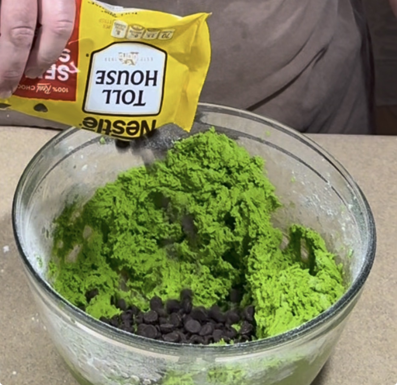 A hand pours a bag of Nestlé Toll House semi-sweet chocolate chips into a glass bowl of bright green cookie dough.
