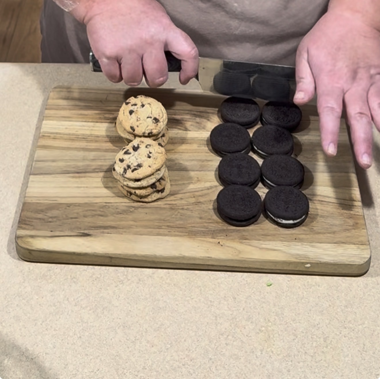 A person uses a knife to cut chocolate sandwich cookies on a wooden cutting board next to a stack of chocolate chip cookies.