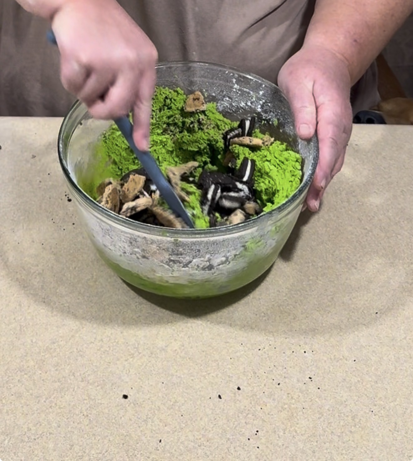 A person uses a spatula to fold chopped sandwich cookies into a bowl of bright green cookie dough.