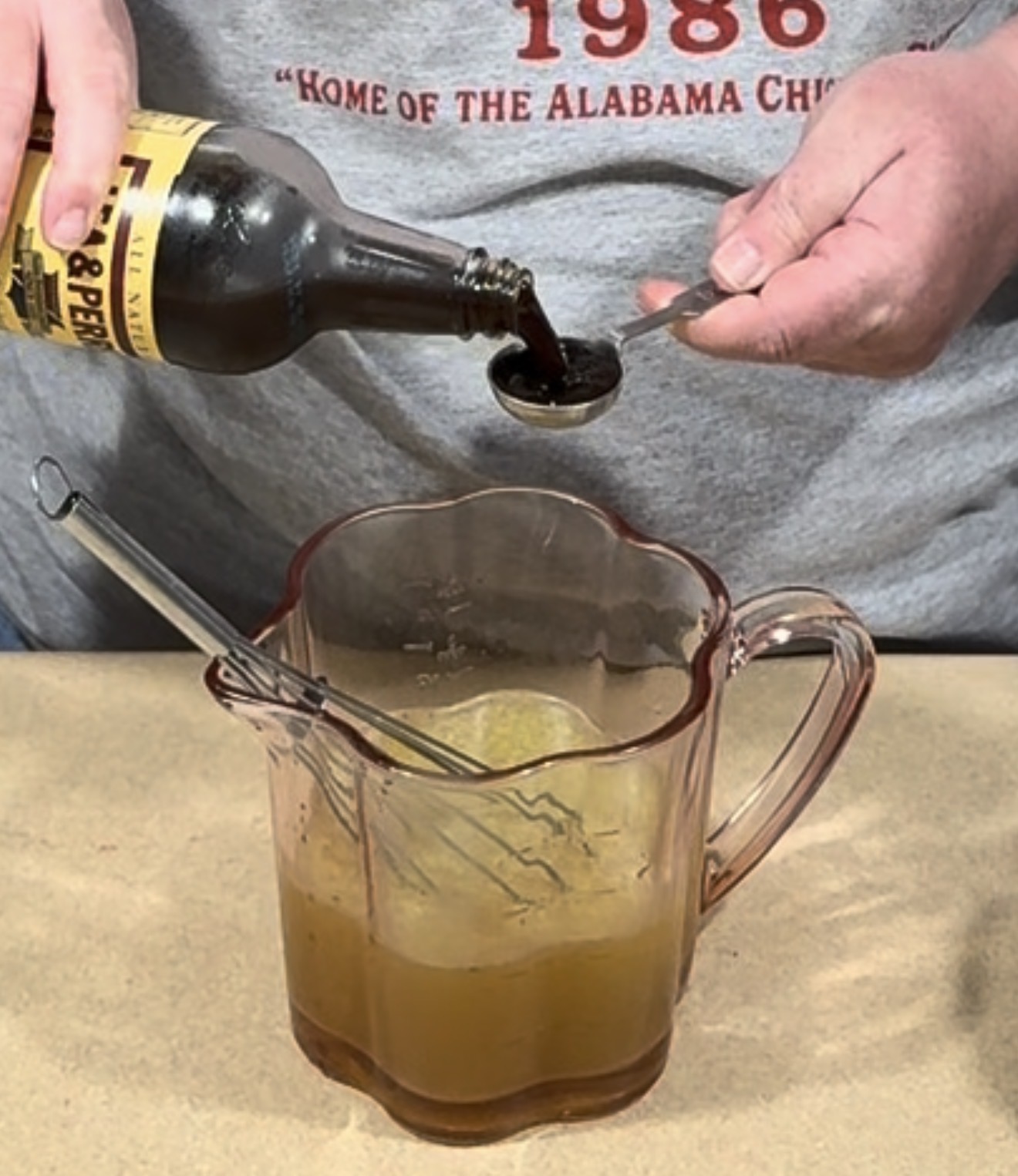 A person pouring Worcestershire sauce from a bottle into a measuring spoon above a glass pitcher containing a golden liquid.