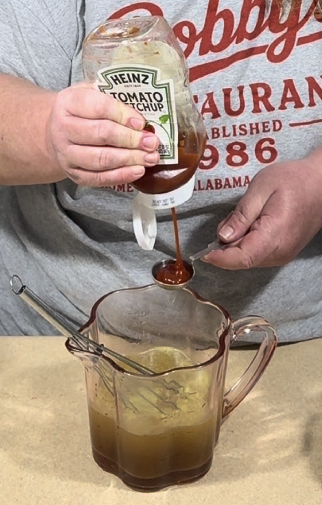 A person pours ketchup from a bottle into a measuring spoon held over a mixing pitcher.
