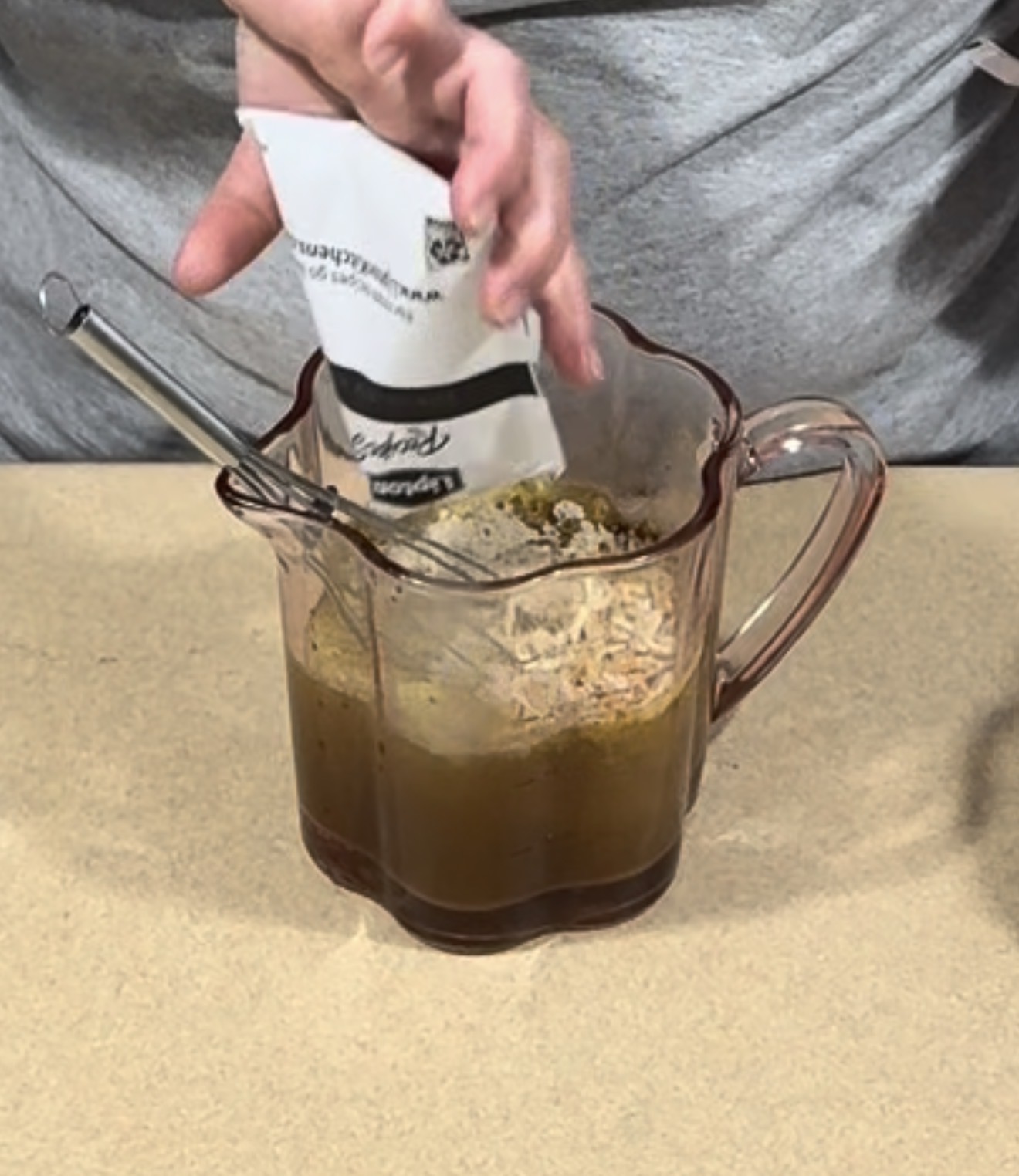 A person pours a packet of Lipton onion soup mix into a glass pitcher containing liquid and a metal whisk.