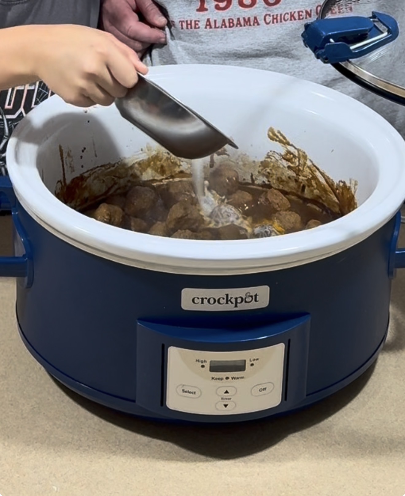 A person uses a metal scoop to pour a white liquid into a blue Crockpot filled with meatballs in sauce.
