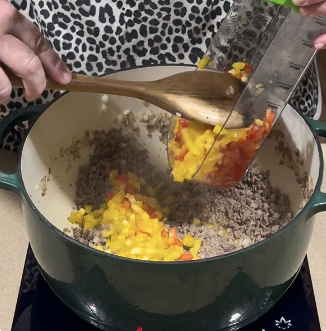 A person uses a wooden spoon to guide chopped yellow and red bell peppers from a measuring cup into a large pot containing browned ground meat.