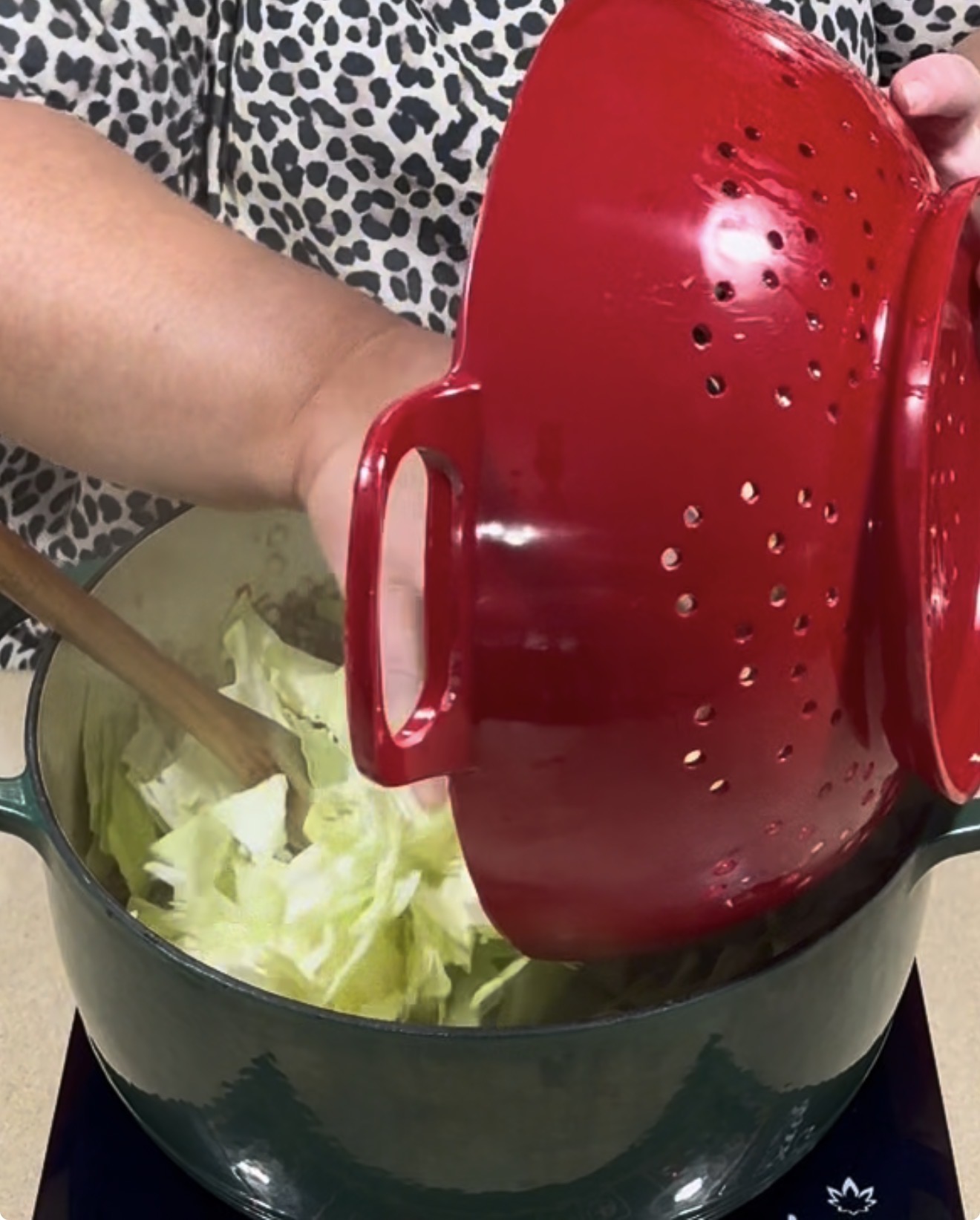 A person pours chopped cabbage from a red colander into a large green pot.