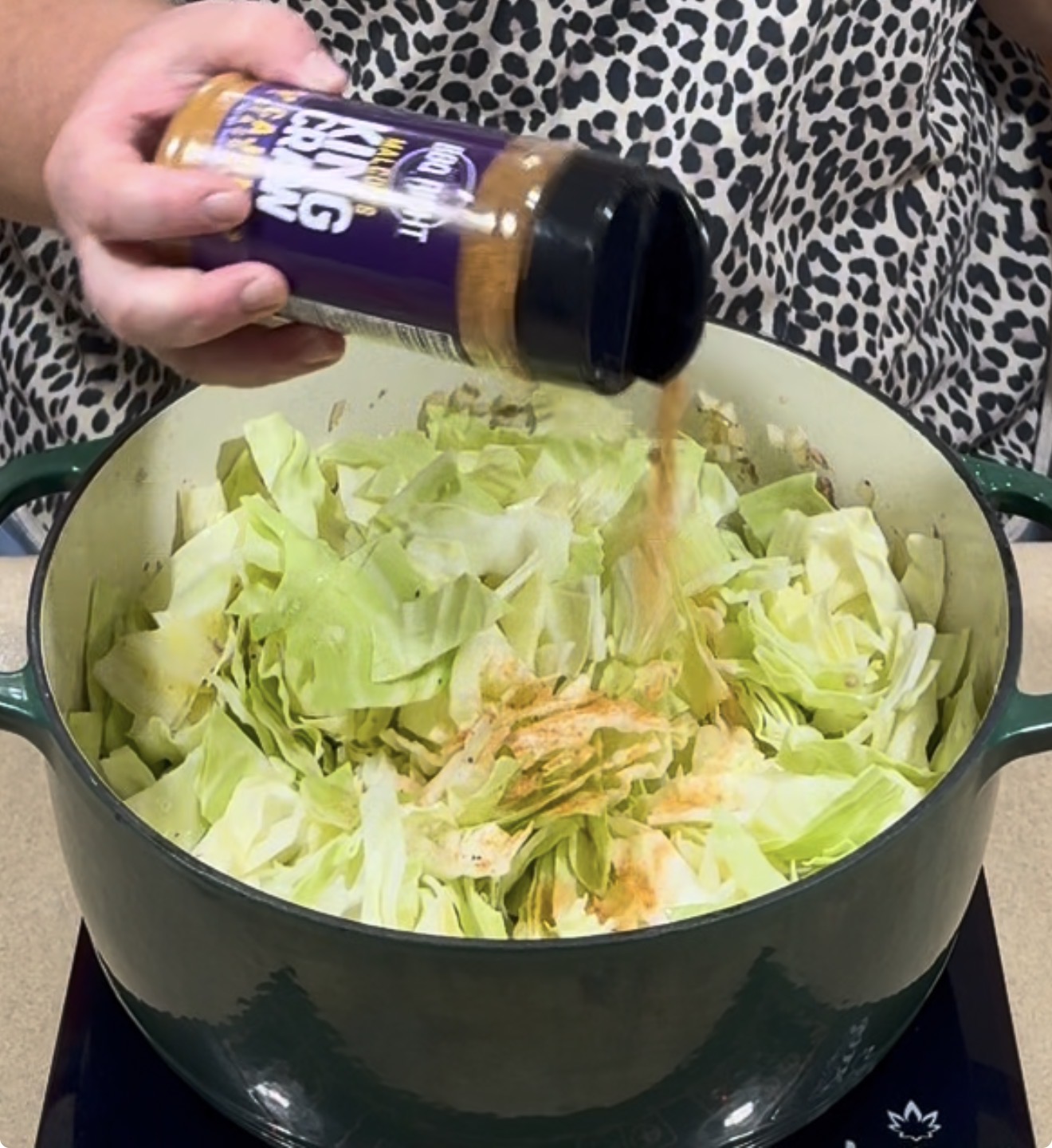 A hand sprinkles seasoning from a bottle labeled King Craw onto chopped cabbage in a large green pot.