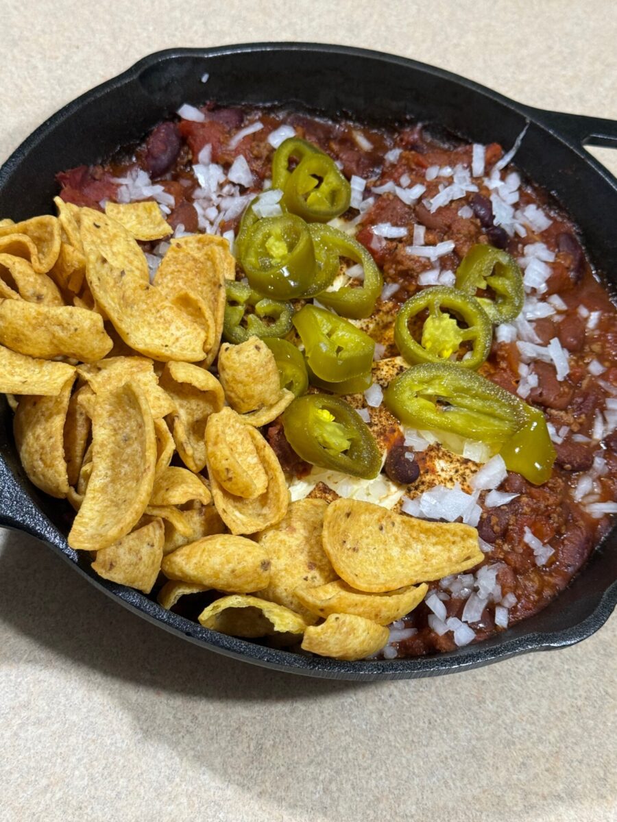 Cast iron skillet filled with chili topped with corn chips, sliced jalapeños, and diced white onions.