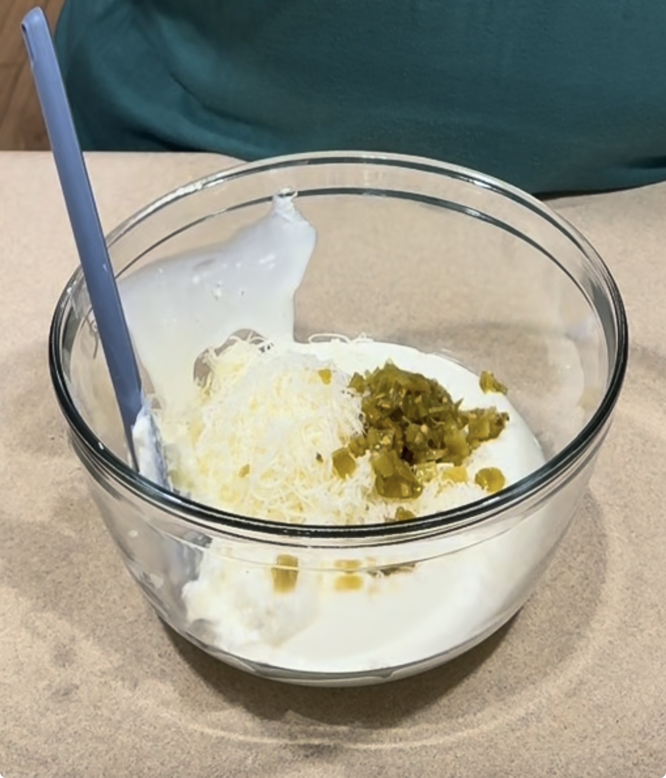 A glass bowl containing sour cream, shredded cheese, and chopped jalapeños, with a blue spatula resting inside.