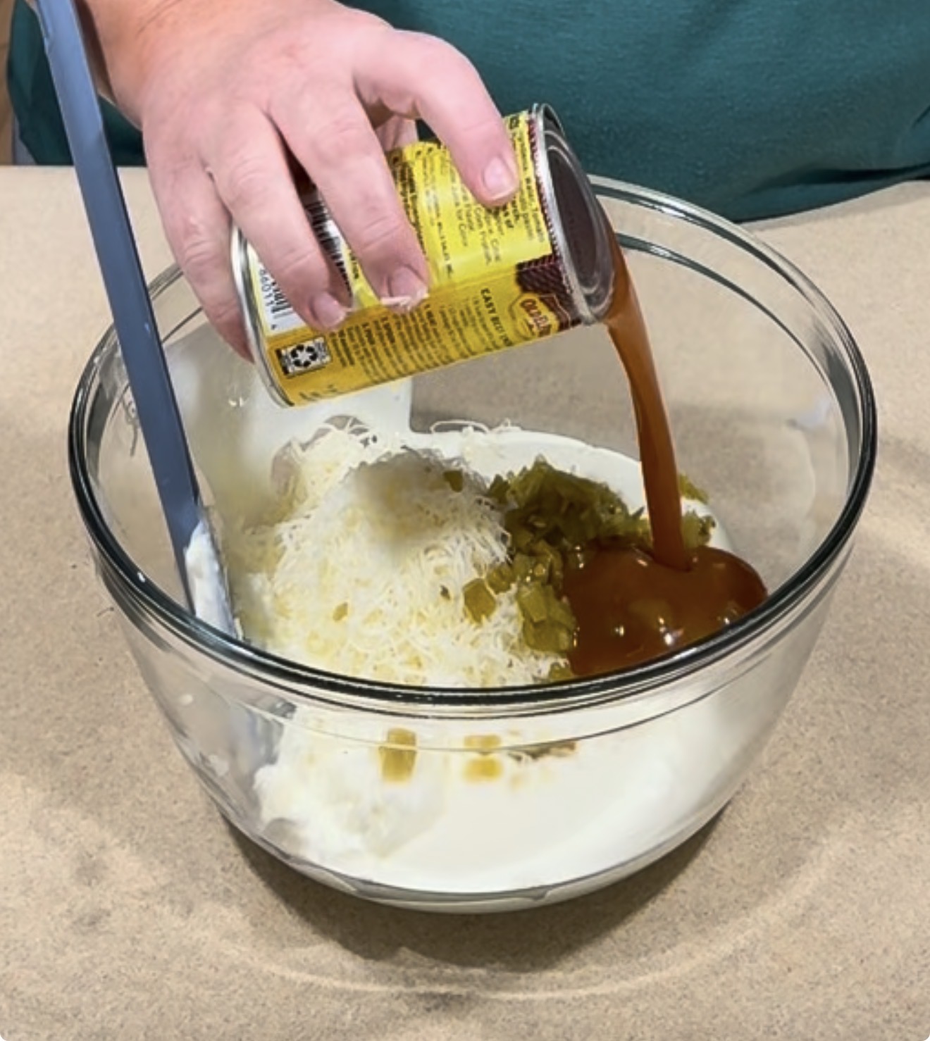 A hand pours a can of dark sauce into a glass bowl containing shredded cheese, chopped jalapeños, and a white creamy base.