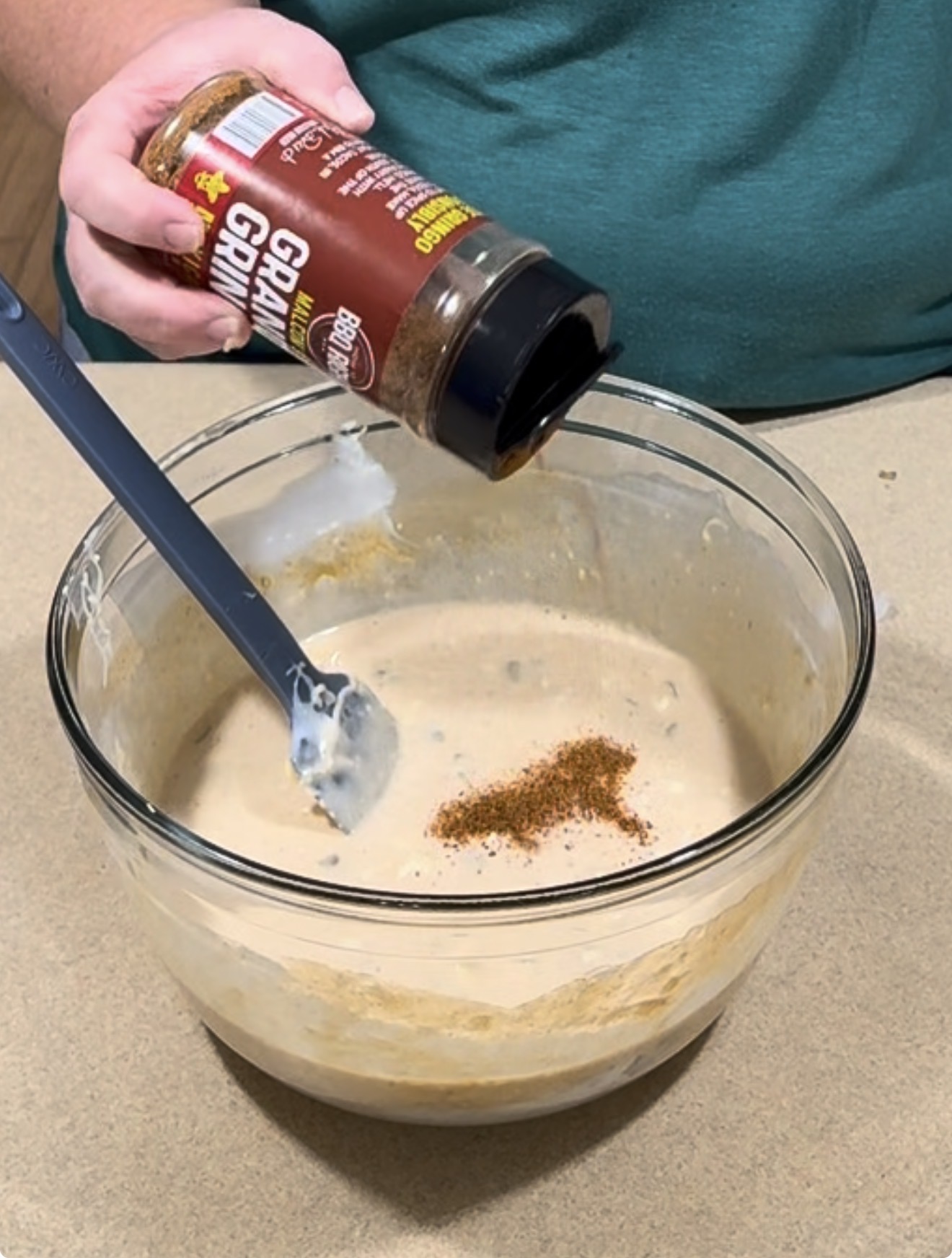 A person sprinkles BBQ seasoning into a glass mixing bowl containing a creamy mixture, with a silicone spatula resting inside.