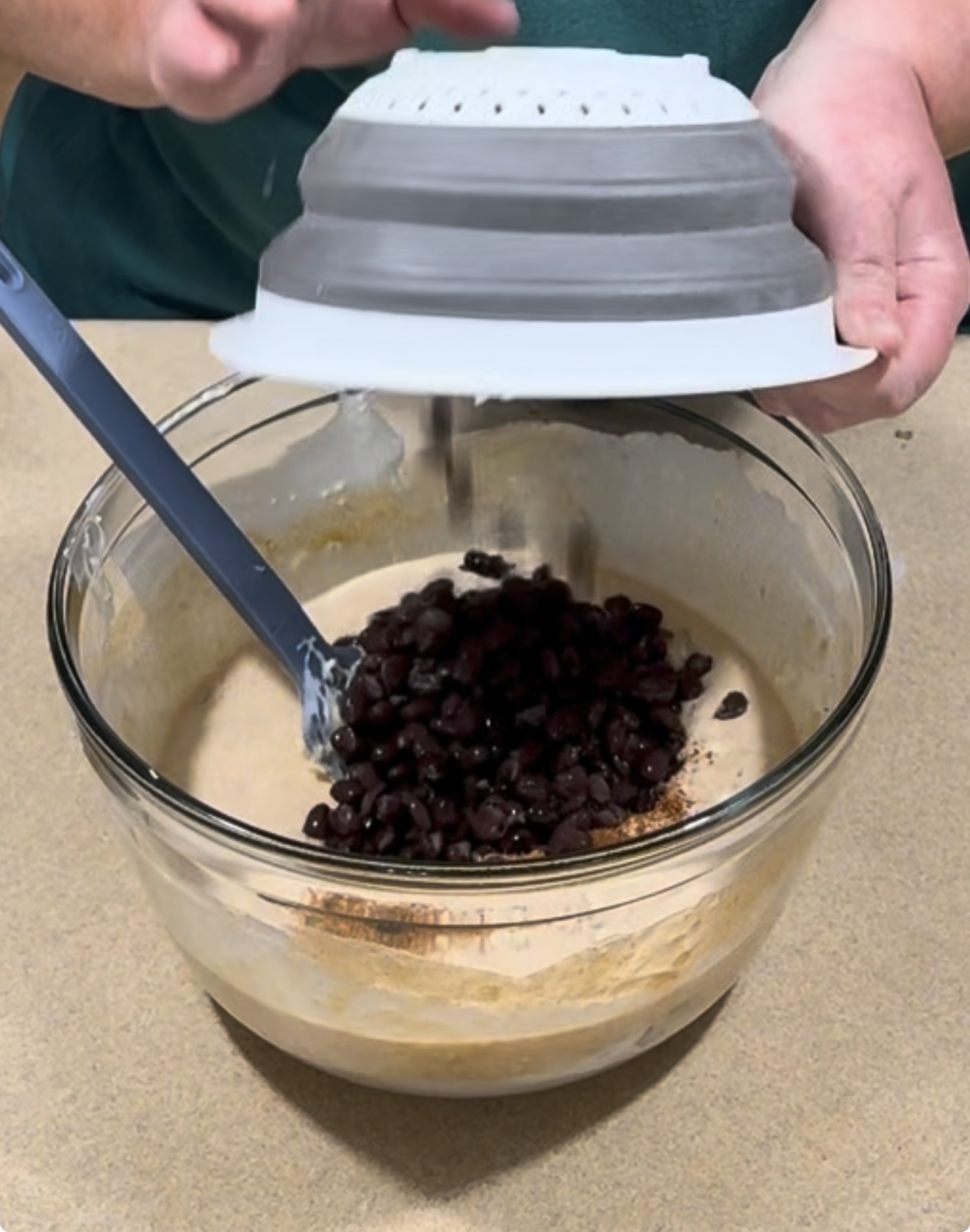 Hands pouring dark chocolate chips from a collapsible colander into a glass mixing bowl containing batter.