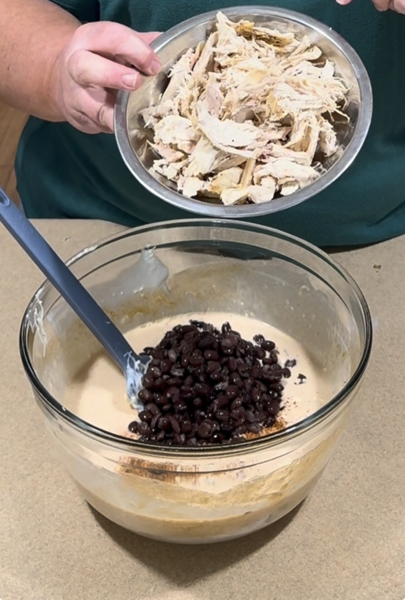 A person holding a bowl of shredded chicken over a glass mixing bowl containing a creamy mixture and black beans.