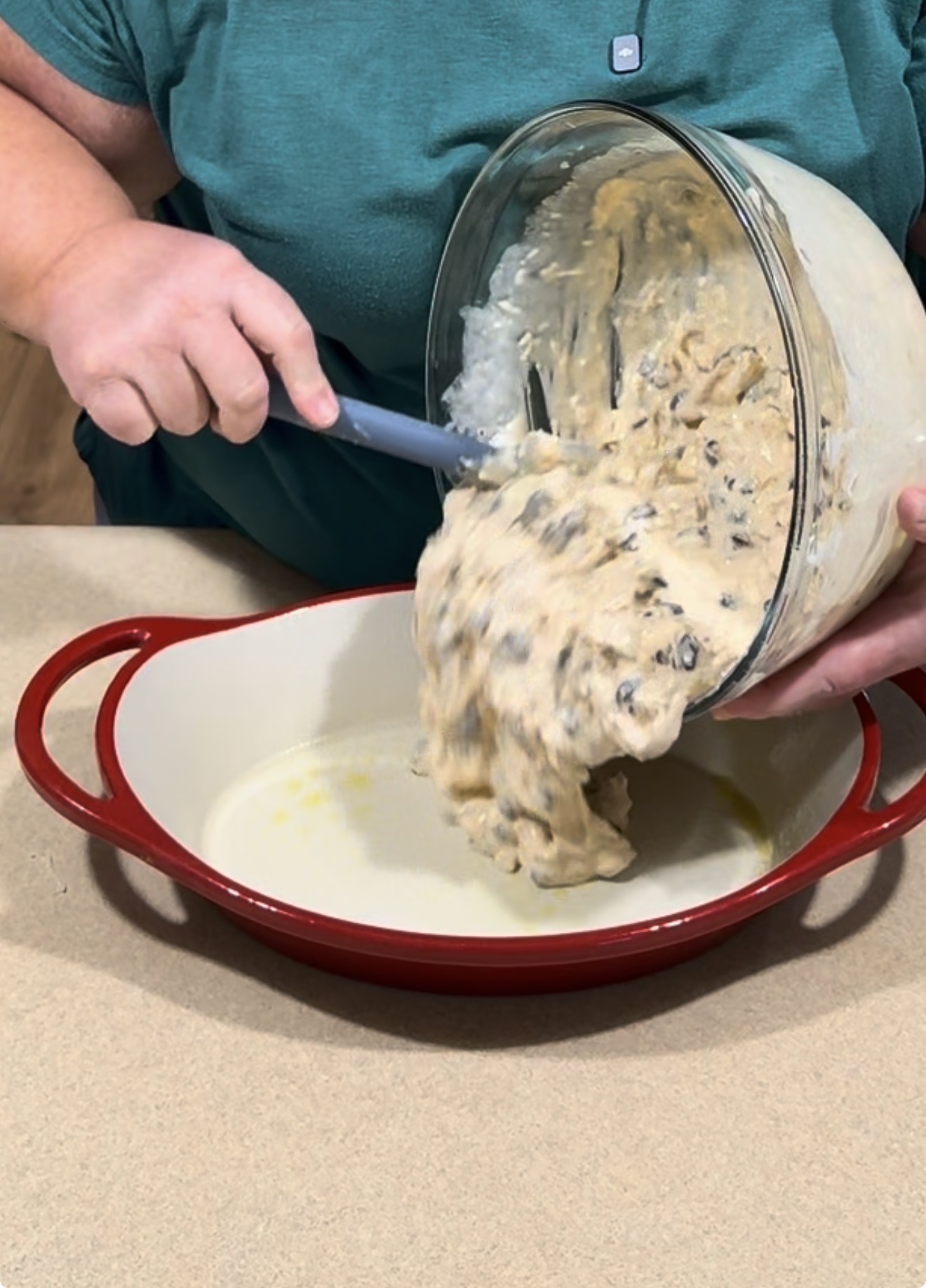 A person pours thick, chocolate chip cookie dough from a glass mixing bowl into a red ceramic baking dish.