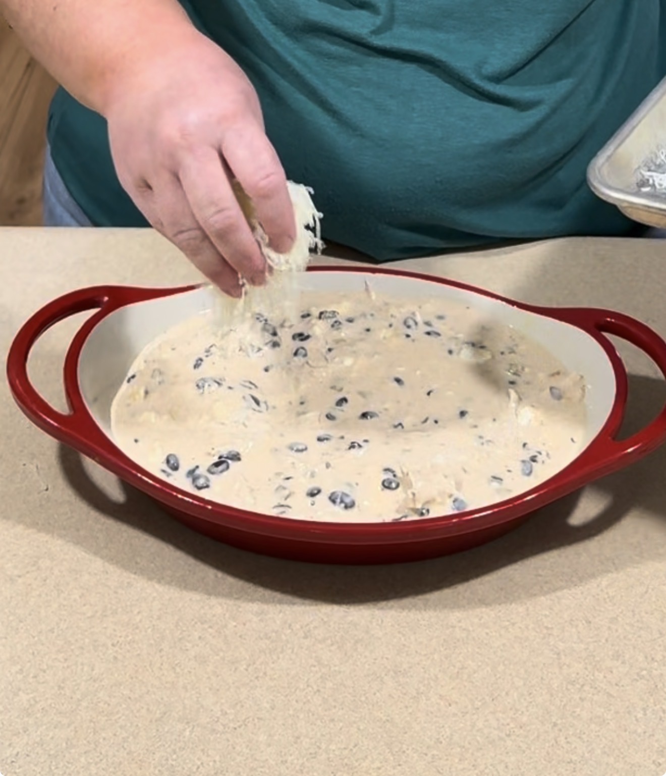 A person sprinkles shredded cheese over a creamy bean dip in a red baking dish.