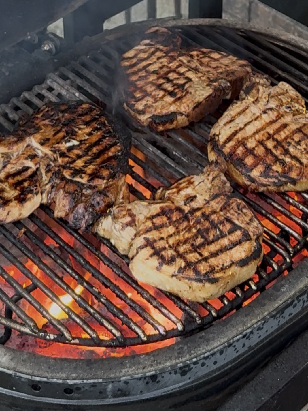 Four thick steaks with char marks cooking on a grill over hot coals.