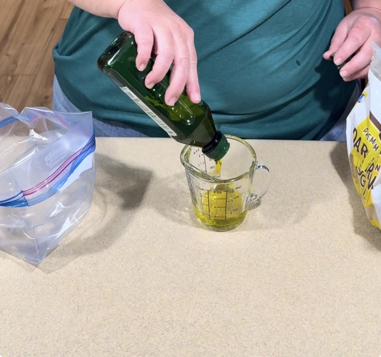 A person pours olive oil from a green bottle into a glass measuring cup on a kitchen counter.