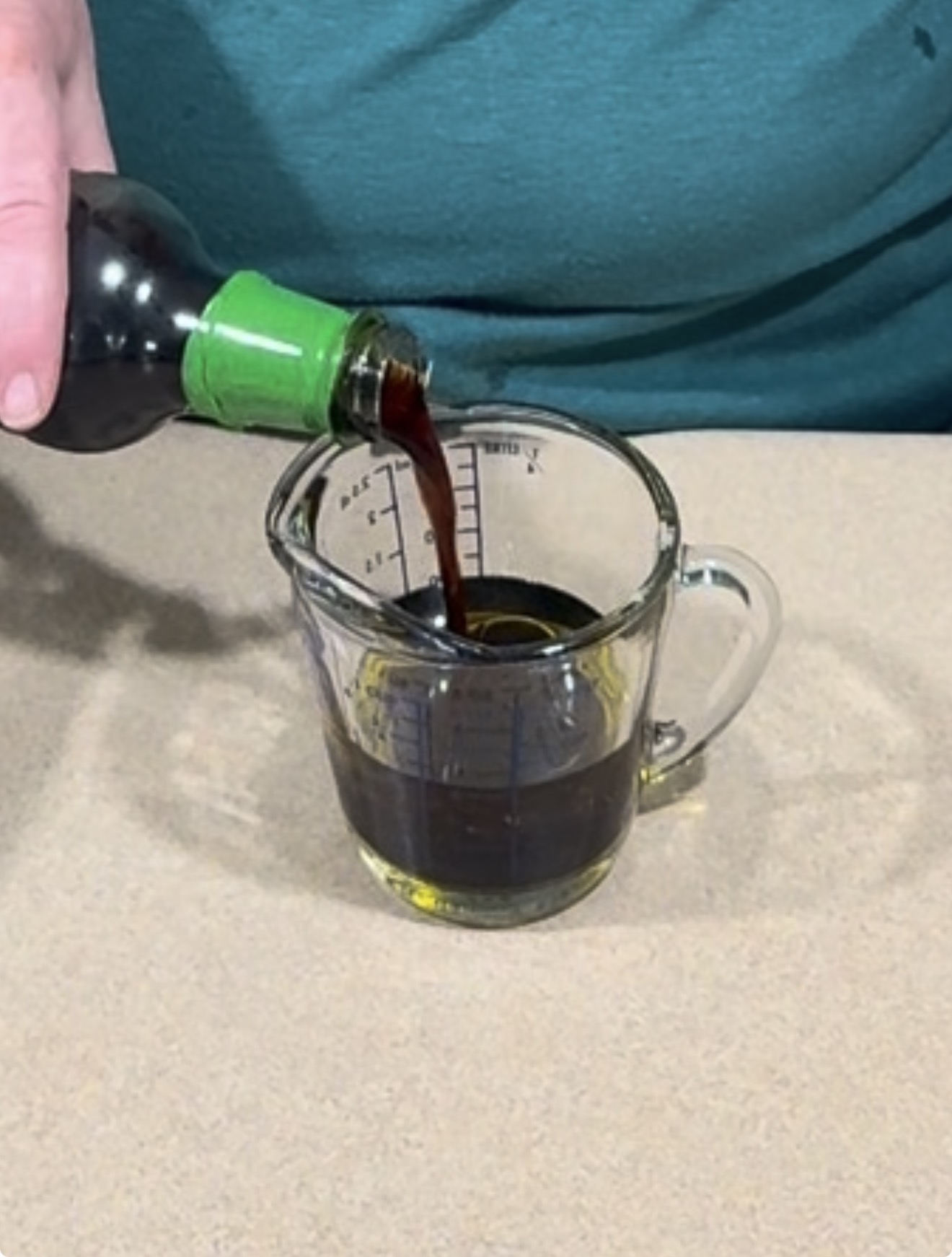 Dark liquid being poured from a glass bottle into a measuring cup already containing a small amount of liquid.