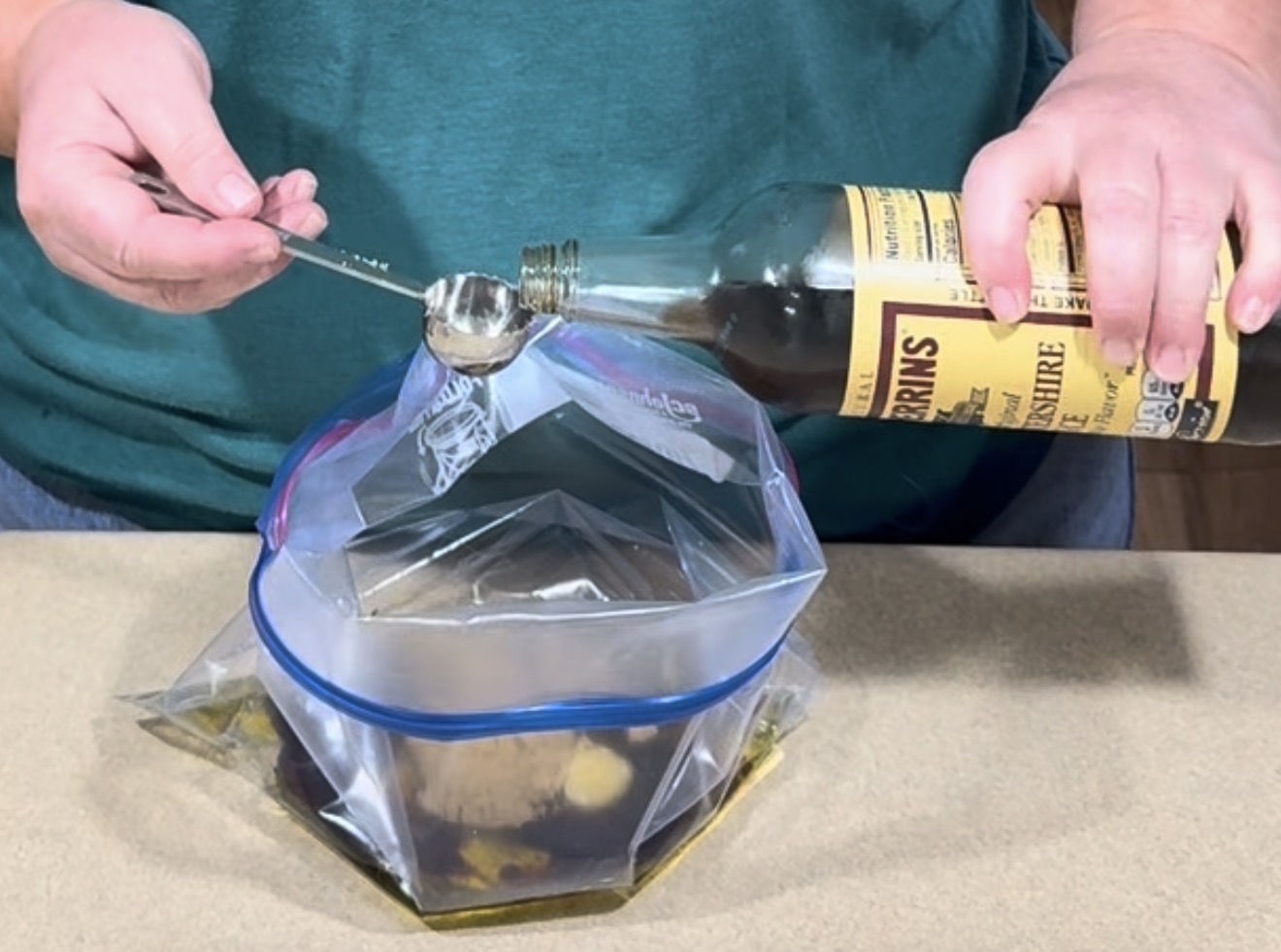 A person pours Worcestershire sauce from a bottle into a plastic storage bag held open on a counter.
