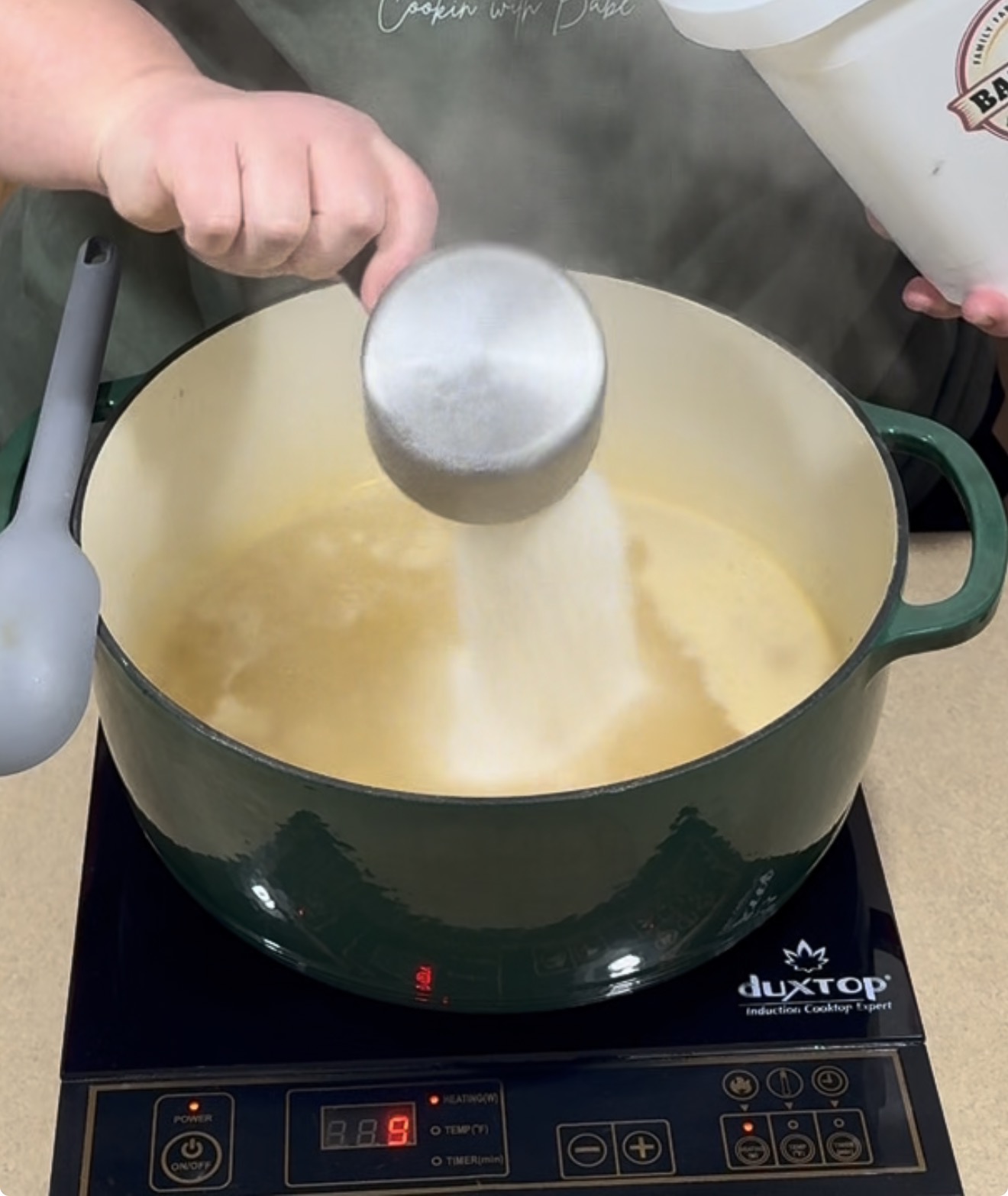 A person pours white powder from a measuring cup into a steaming pot of liquid on an induction cooktop.