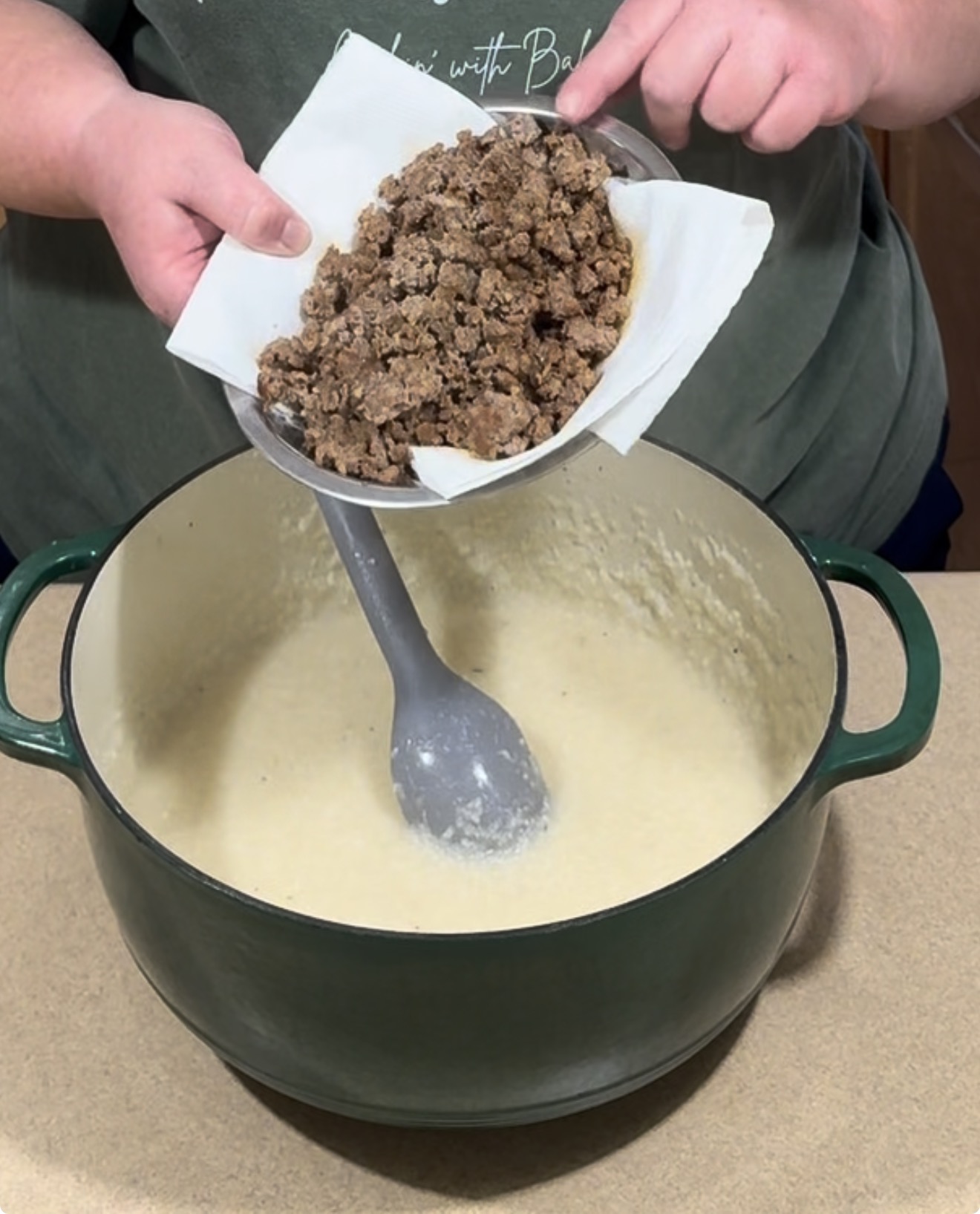 A person pours cooked ground meat from a paper-lined bowl into a large pot of creamy sauce.