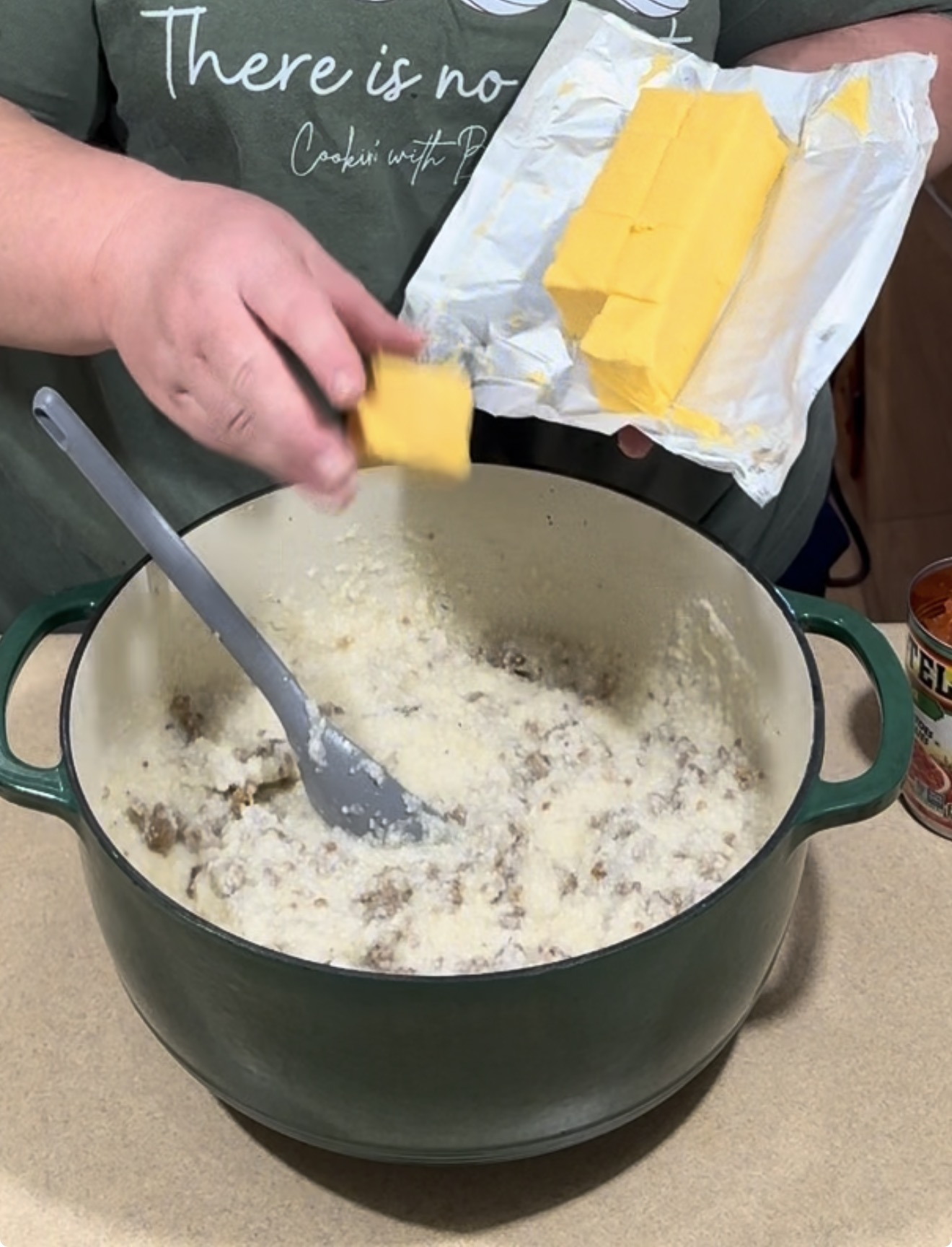 A person dropping a cube of butter into a green pot filled with a creamy, meat-based mixture.