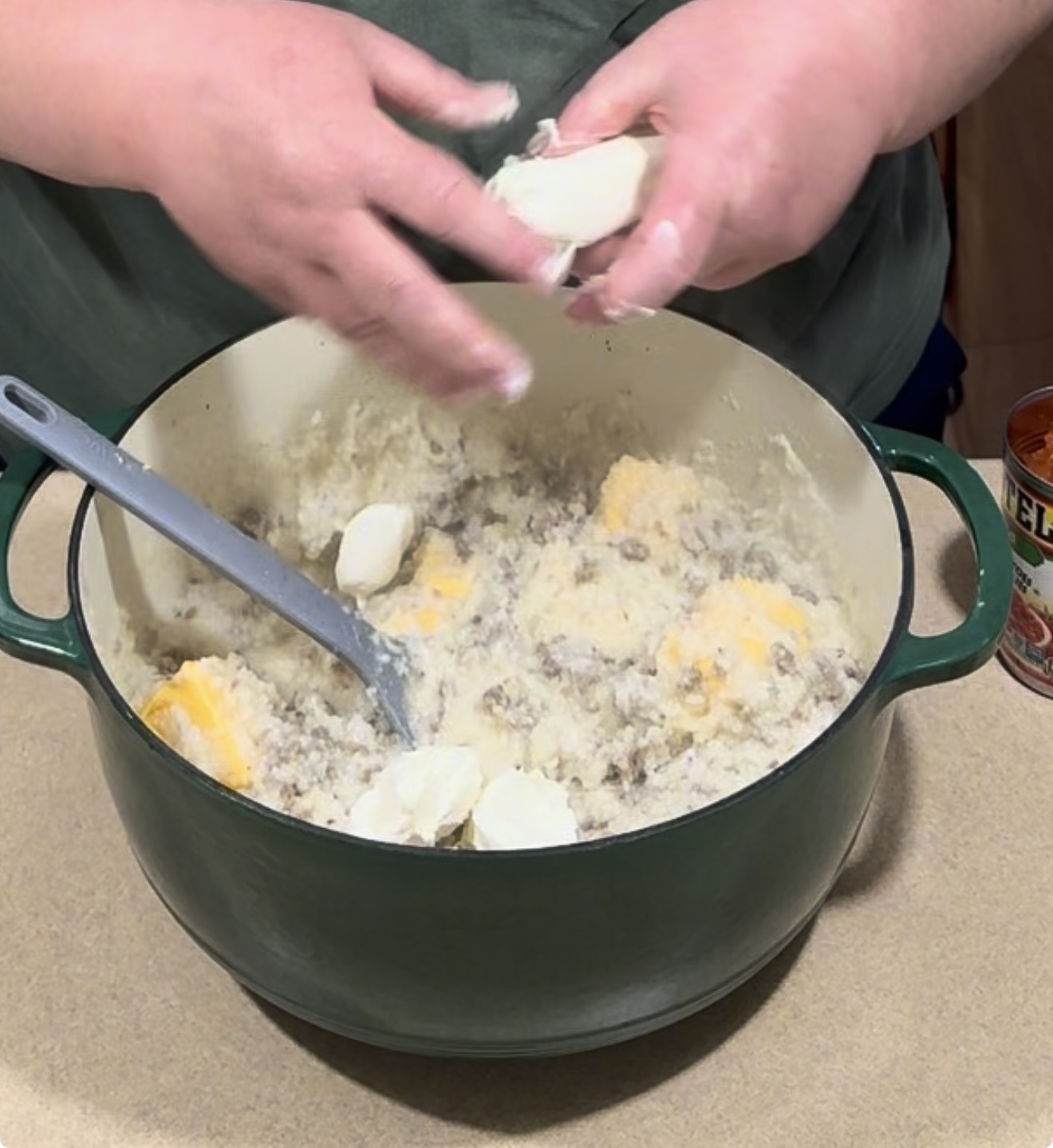Hands add pieces of cheese to a large pot containing a mixture of ground meat and cooked grits.