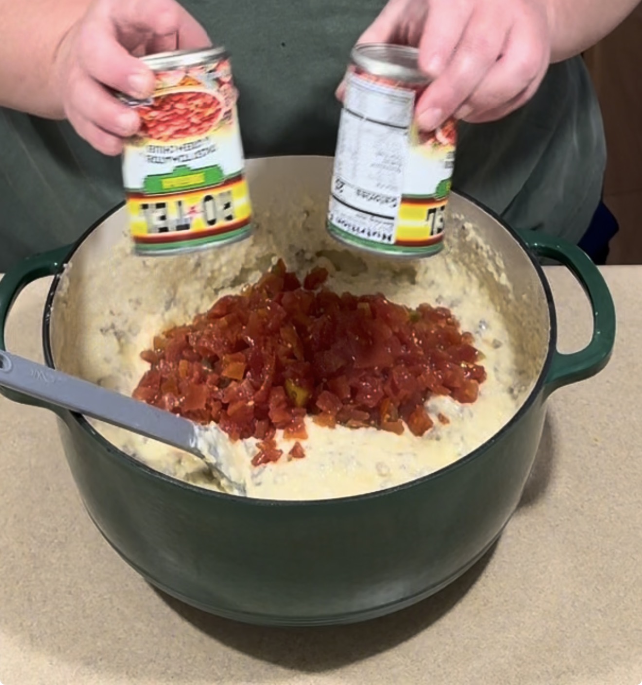 A person pours two cans of Ro-Tel diced tomatoes and green chilies into a large pot of creamy dip.