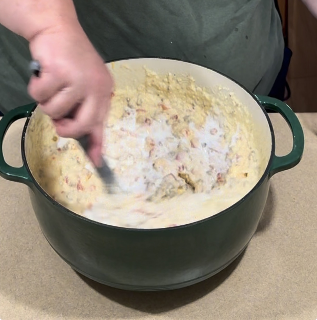 A person uses a spoon to stir a creamy, chunky mixture in a large green pot.