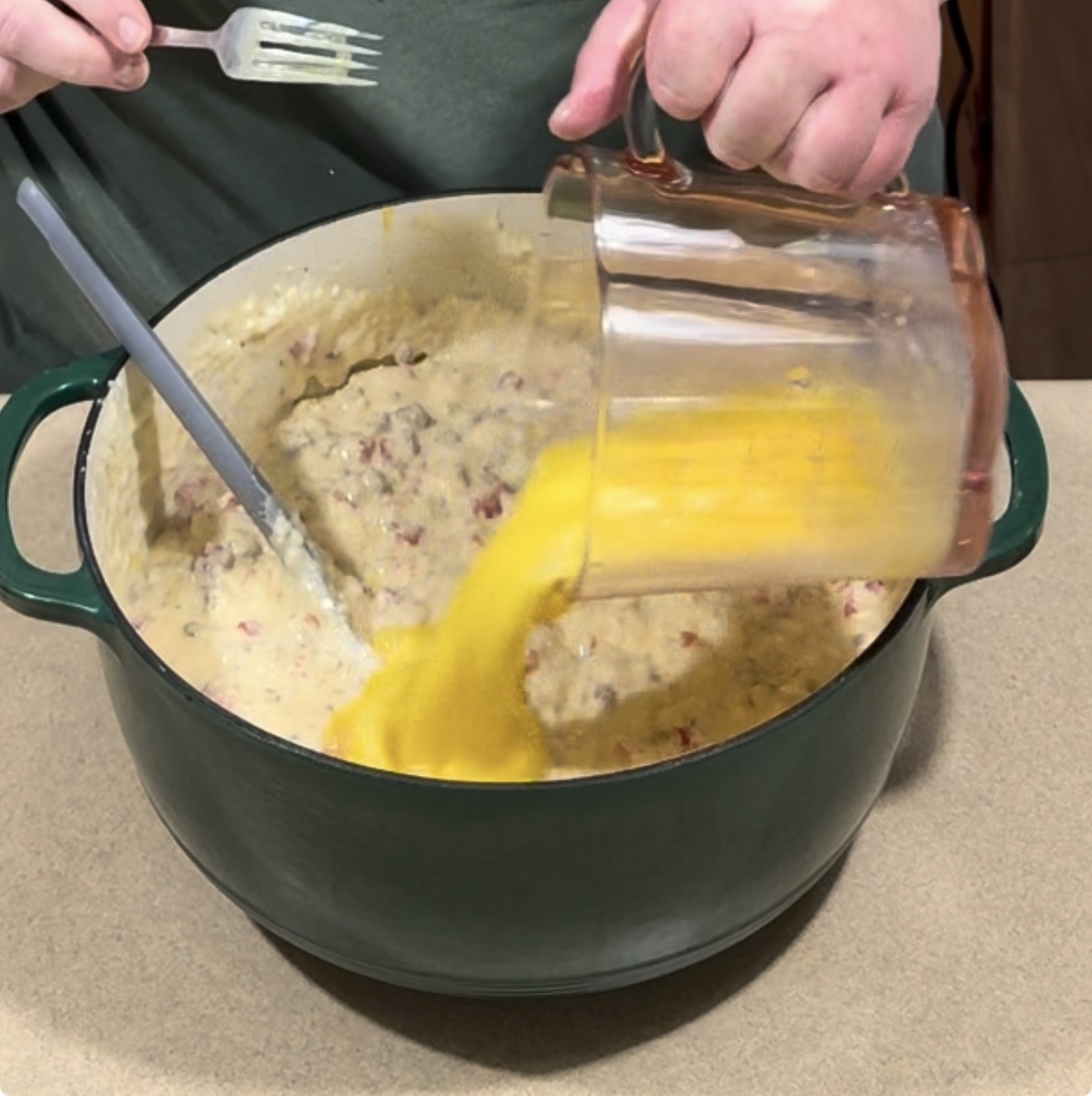 A person pours beaten eggs from a glass measuring cup into a large green pot containing a creamy, vegetable-based mixture.