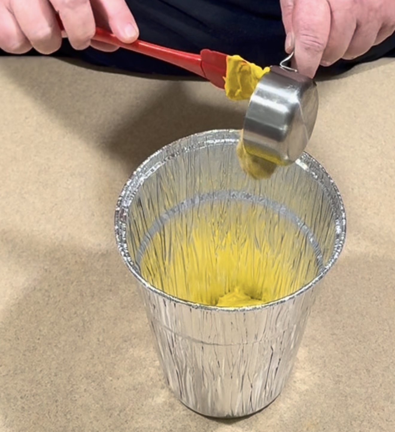 Hands using a red spatula to scrape yellow paste from a metal measuring cup into a foil container.