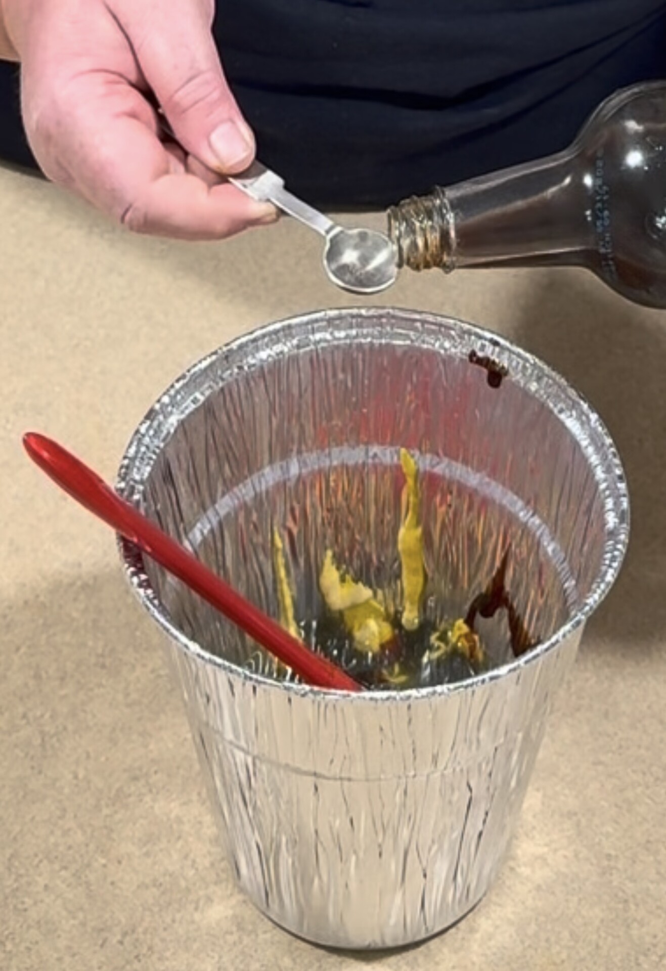 A hand pours dark liquid from a small measuring spoon into a foil cup containing yellow mustard and a red stirring stick.