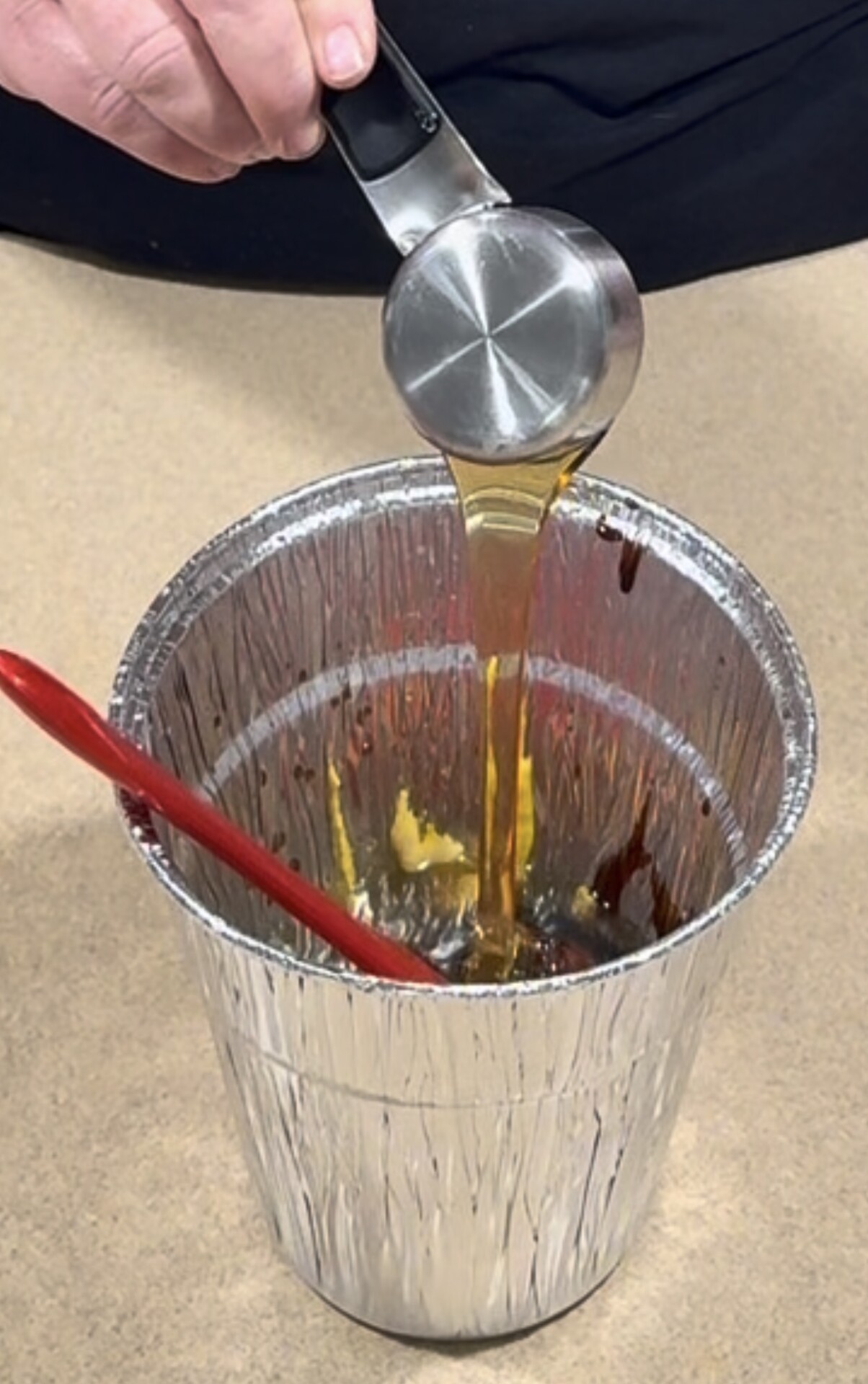 Honey pours from a metal measuring cup into a foil container holding other ingredients.