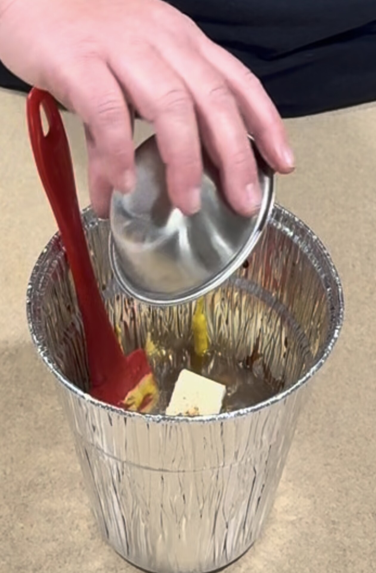 A hand holding a small metal bowl pours yellow liquid into a foil container holding a stick of butter and a red silicone spatula.