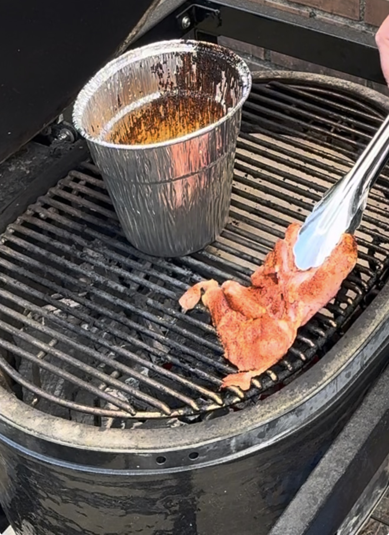 Tongs placing a piece of seasoned raw meat onto a grill grate next to a disposable foil grease cup.