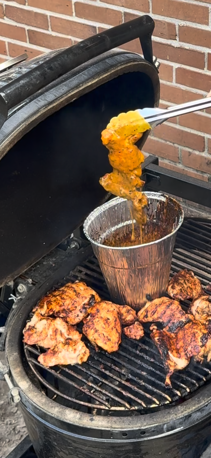 A hand uses tongs to dip a piece of marinated chicken into a metal container on a grill filled with other pieces of charred chicken.