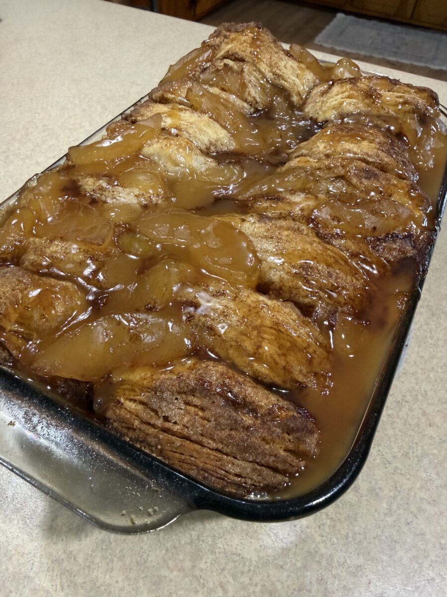 A glass baking dish filled with golden-brown cinnamon rolls topped with glossy apple pie filling and syrup on a tan countertop.