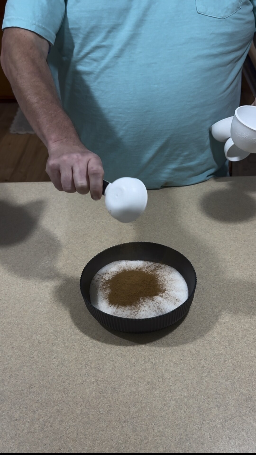 A person holds a white measuring cup over a black bowl filled with sugar and cinnamon on a beige countertop.