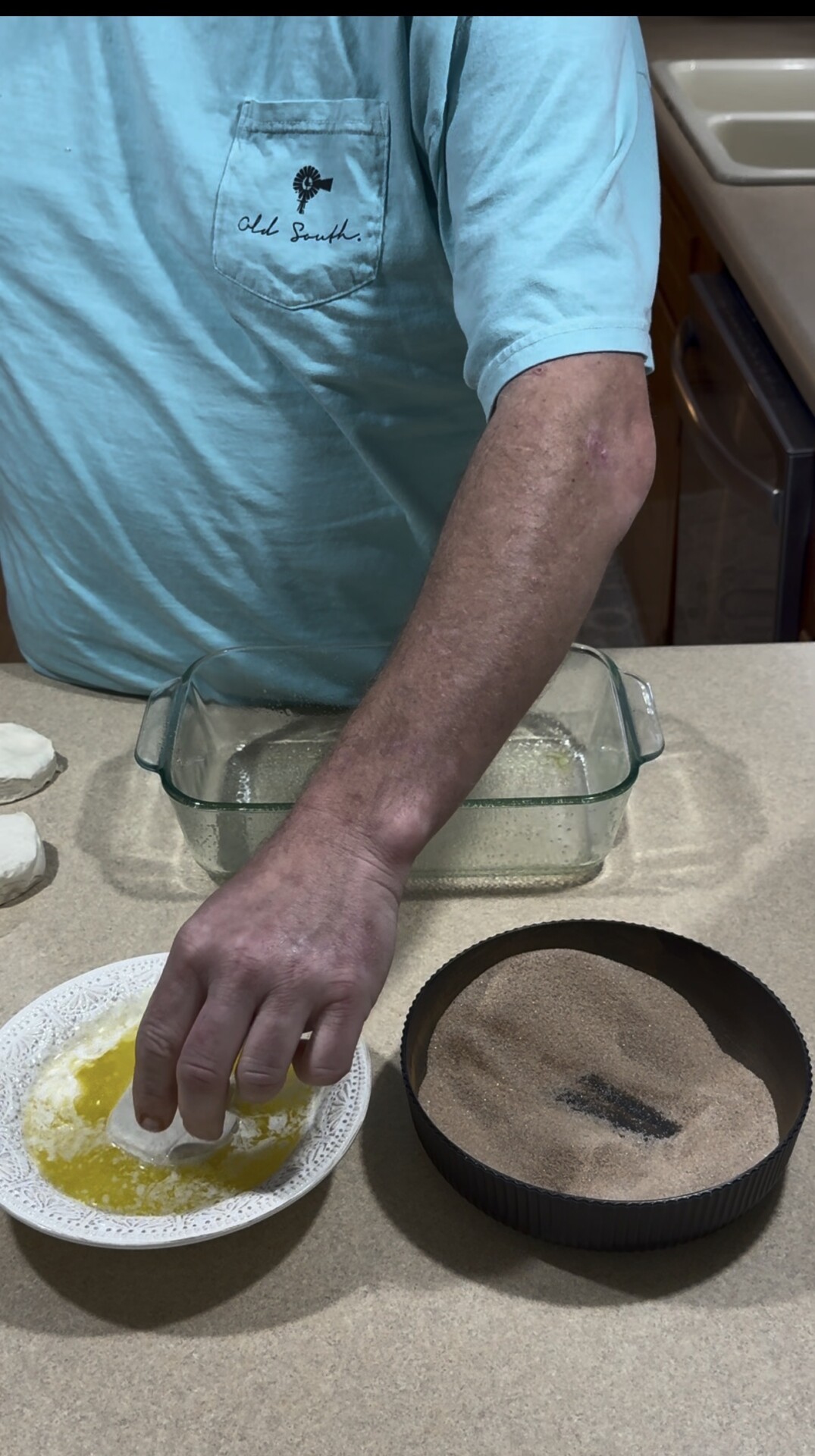 A person dips dough into melted butter and cinnamon sugar, with a glass baking dish nearby on a kitchen counter.