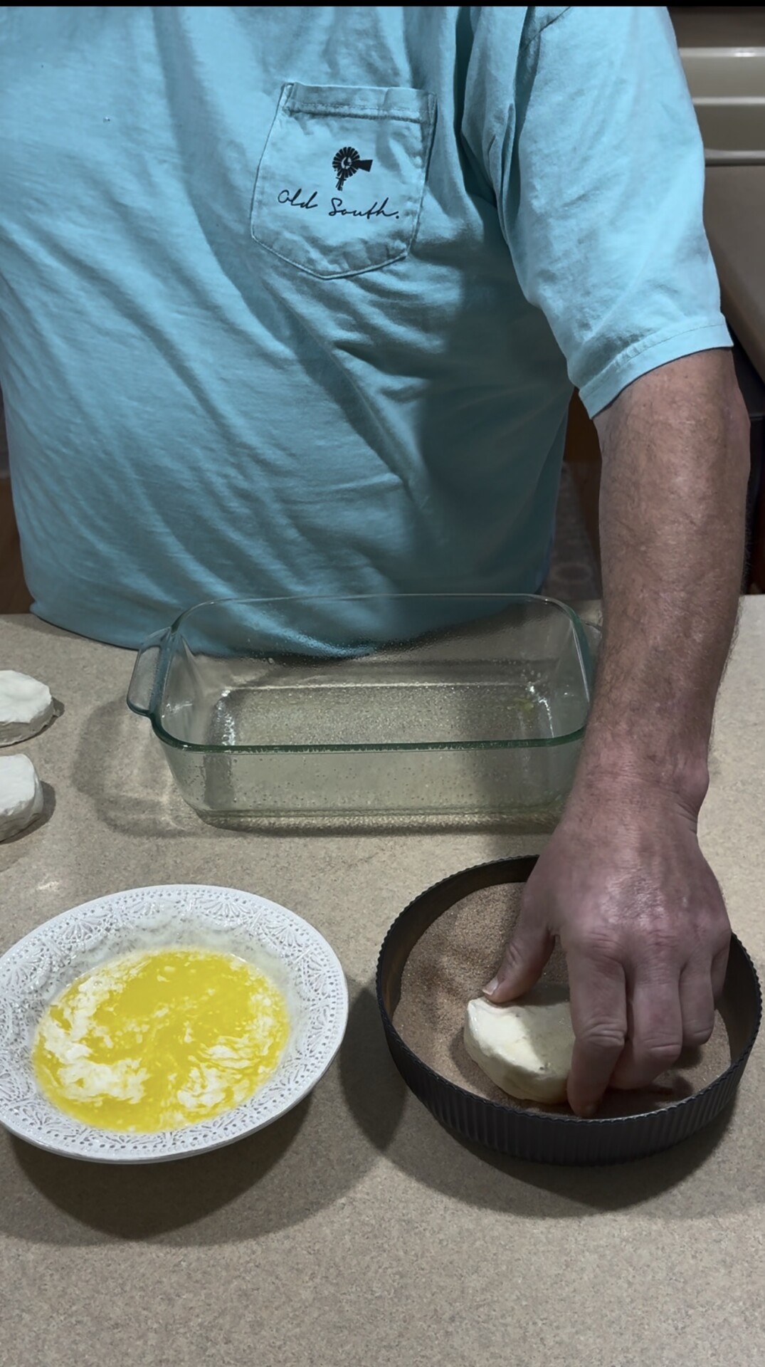 A person in a blue shirt dips dough into a round dish next to melted butter in a bowl and a glass baking dish on a kitchen counter.