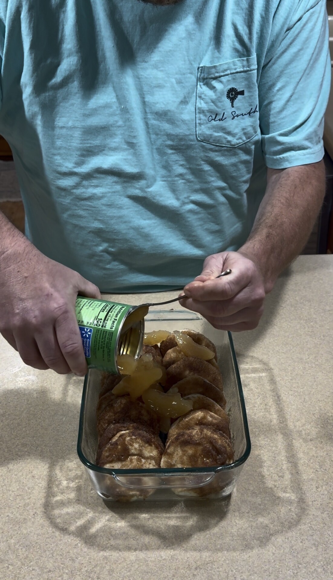 A person in a blue shirt pours canned sliced peaches over cinnamon rolls arranged in a glass baking dish on a kitchen counter.